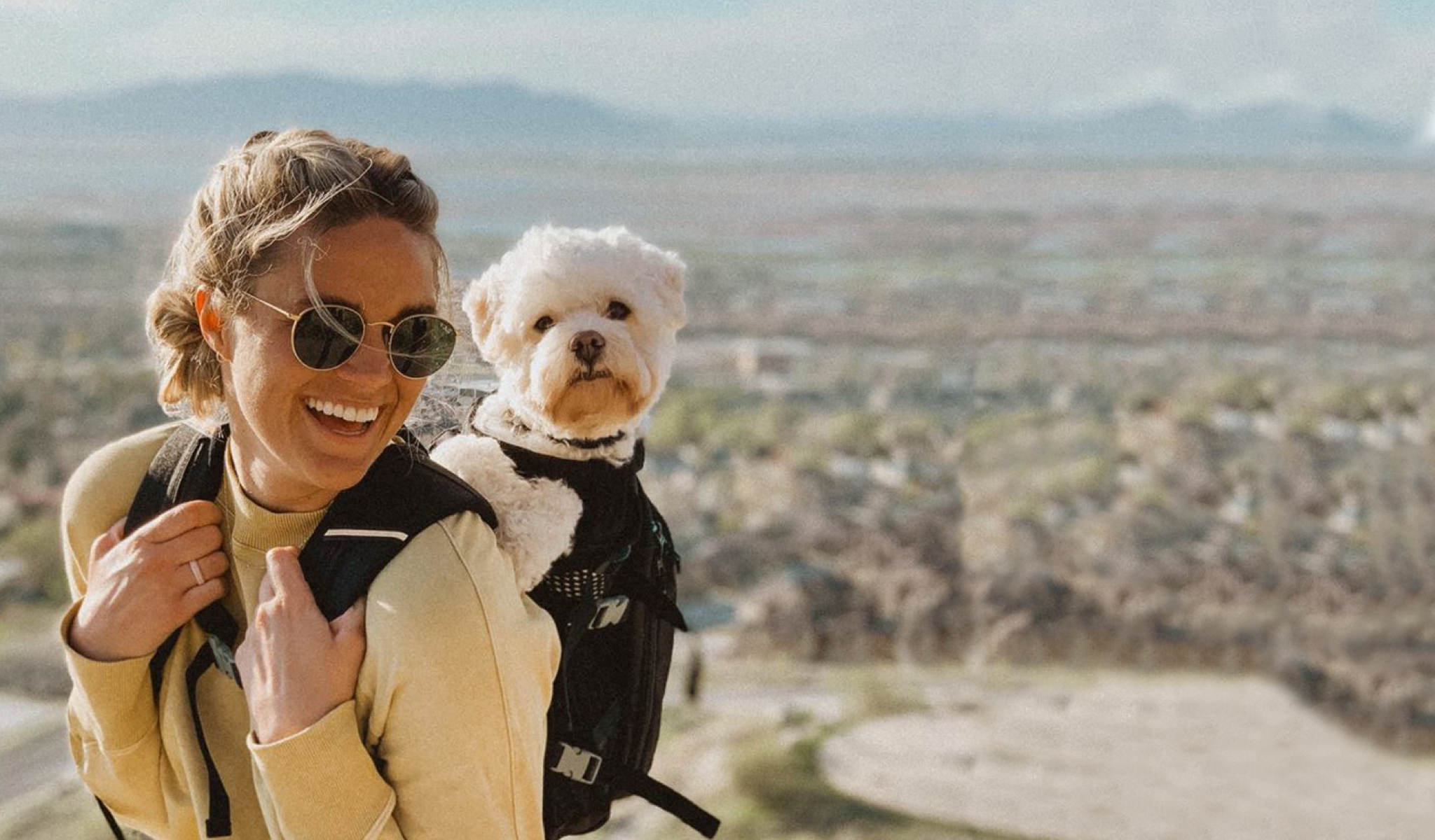 A woman in sunglasses enjoys time on a mountainside with her dog on her back in a K9 Sport Sack Plus 2 backpack carrier